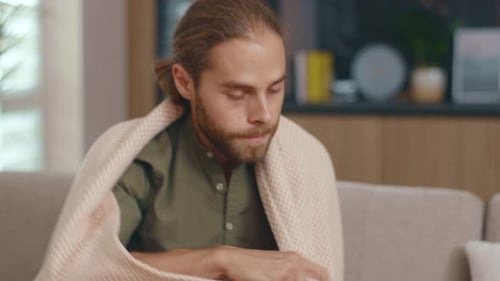 Jeune homme hipster malade souffrant de froid et de hautes températures portant une couverture chaude assis sur un canapé