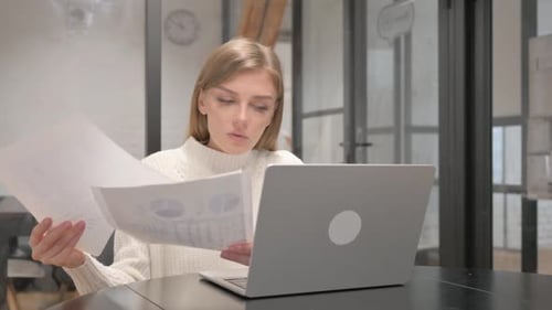 Aufgeregte kreative junge Frau feiert, während sie an Dokumenten und Laptop arbeitet