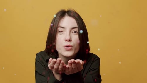 Woman Blowing Confetti Toward Camera on Yellow Background