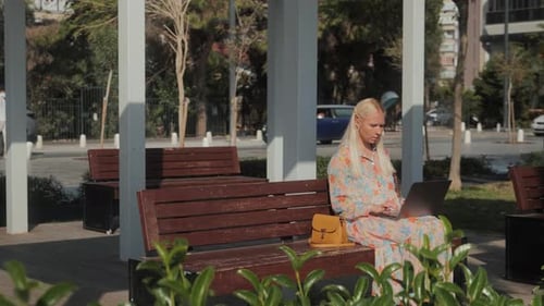 Young Beautiful Blonde Woman is Sitting on a Park Bench and Working on a Laptop