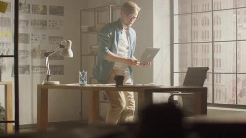 Creative Designer Walks around His Studio, Sits on His Desk Holds Laptop on the Knees while Working