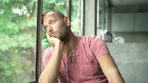 Melancholic man sits thoughtfully by window gazing at rainy garden view