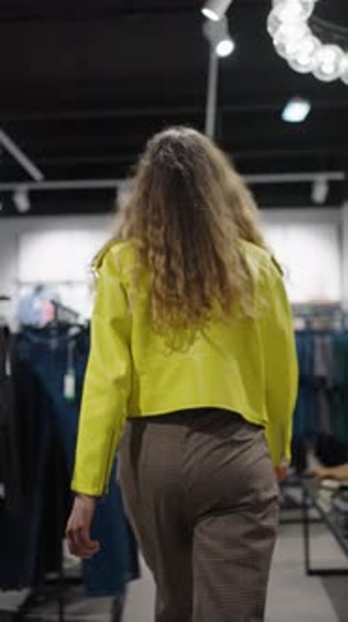 Woman in Yellow Jacket Walks Between Hangers of Clothes in Fashion Store