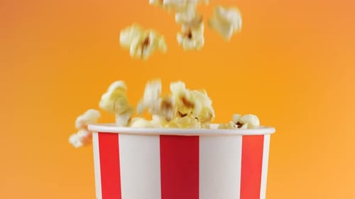 Popcornkörner, die einzeln auf pfirsichfarbenem Hintergrund in einen rot-weiß gestreiften Eimer fallen