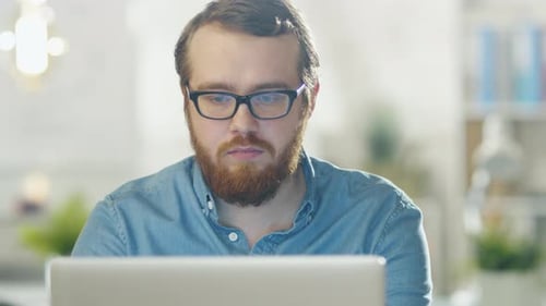 Portrait of a Bearded Young Man Wearing Glasses Sitting in His Office Working on a Computer. Comput