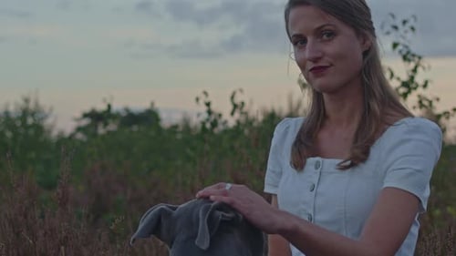 Portrait of attractive young woman petting the head of her pitbull in a heather field at dusk and lo