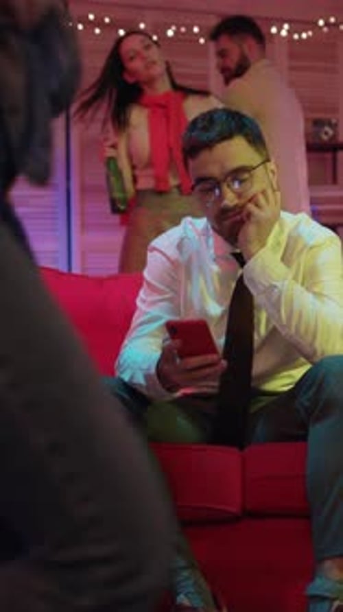 A Distracted Man Sitting on His Couch During a Lively Social Gathering with Friends