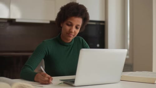 Focused Young Female Black Freelancer Using Laptop Writing Notes Sitting at Home Office Desk Smiling