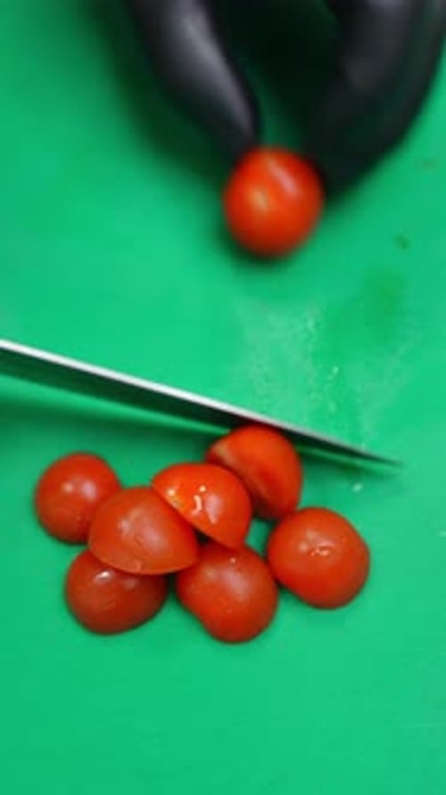 Mit Handschuhen Cherrytomaten auf einem grünen Schneidebrett in Scheiben schneiden und dann in eine Metallschüssel geben