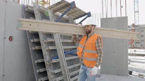 Builder man in hardhat and vest carrying timber on building site in his working day