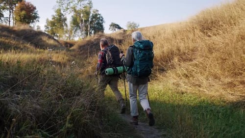 Un anciano y una mujer con bastones caminan entre colinas con hierba seca