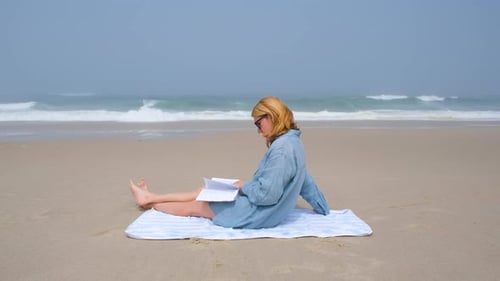 Eine junge Frau sitzt auf einem Handtuch am Atlantik und liest ein Buch an einem ruhigen Sandstrand