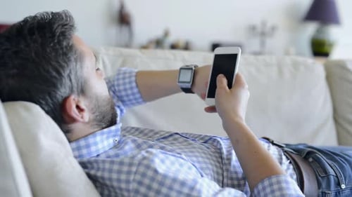 Man relaxing on couch using phone and smartwatch