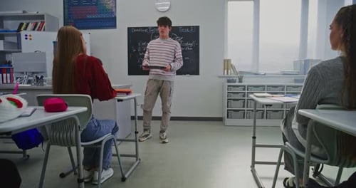 Young Male Student Stands at Front Classroom Holding Digital Tablet While Delivering Report