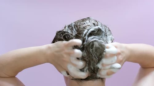 A woman washes her hair with shampoo on lilac background, rear view.
