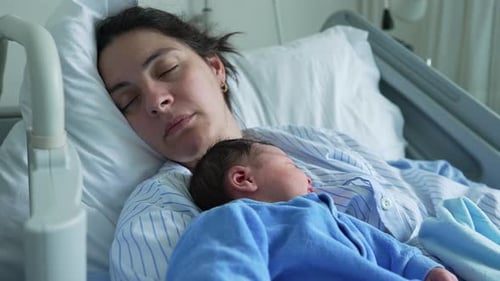 Mother and newborn baby sleeping together in a hospital bed, both in matching blue outfits,