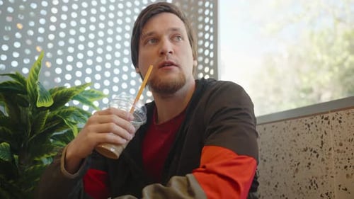 Man in a Red and Black Jacket Sitting at a Restaurant Table Sipping a Cold Beverage Through a Straw