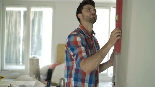 Junger Mann mit Bauplänen überprüft Wand mit Wasserstand im neuen Zuhause der 20er