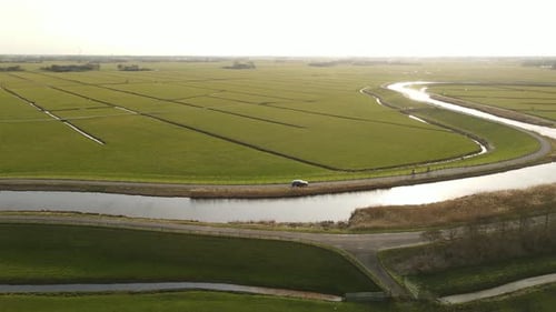 Aerial drone of two cyclists and car driving parallel to river in green farming area