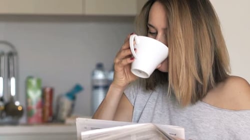 Woman Reading Newspaper and Drinking Coffee in Kitchen