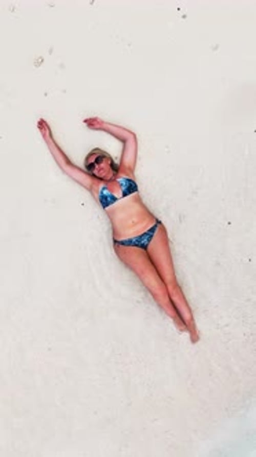 Frau im schwarzen Badeanzug liegt auf nassem Sand, während sanfte Wellen die Küste berühren. Mädchen sonnt sich auf Weiß