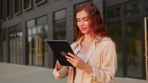 Young Red Haired Woman Holding a Tablet Standing Near Modern Building
