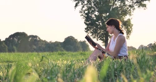 On a sunny day, a beautiful young student girl reads a book in nature (in a park, on a field) agai
