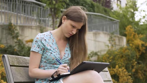 Young Woman Writes in Notebook on Park Bench