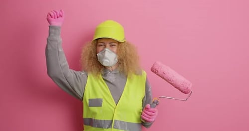 Joyful construction worker woman dances actively with paint roller against pink background