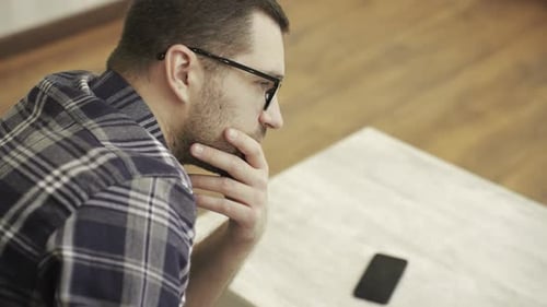 Pensive Man with Glasses Thinking Next to Smartphone
