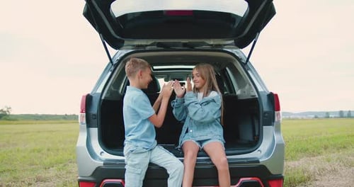 Happy Teen Boy and Girl Which Sitting in Car Trunk Among Field and Playing Funny Games