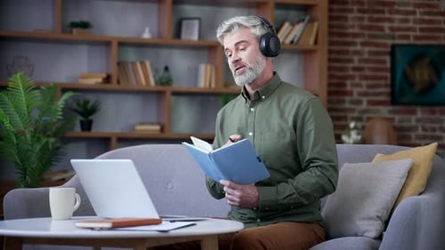 Bearded man with gray hair wearing headset sitting at home office watching online training on laptop