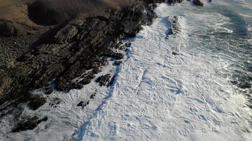 aerial shot of ocean waves hitting rocky shore crashing violently with sun reflecting on the surface