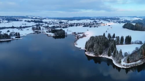Snow blankets the fields and trees around a lake. A small village sits quietly near the water.