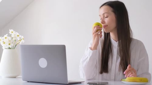 A Brunette Woman Works From Home at the Computer and Eats an Apple