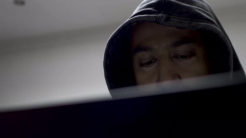 A shot of an anonymous hacker as he works behind a laptop computer, glancing down as he types on the