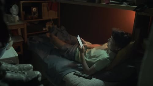 Chinese Man Lying on Bunk with Smartphone in Dark Shoebox Flat