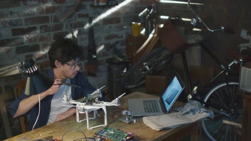 Student is soldering electrical components on a drone in a garage while checking a laptop computer.