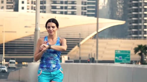 Attractive young woman checks pulse on smartwatch while running in the city slow motion