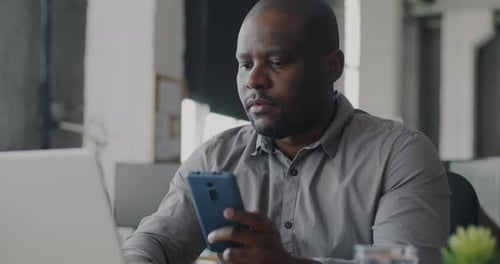 African American Entrepreneur Texting with Smartphone and Typing with Laptop in Coworking Office