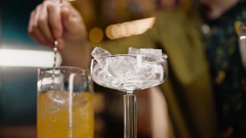 A classic bartender stirs alcohol in a tall glass with a long spoon to prepare a cocktail at the bar