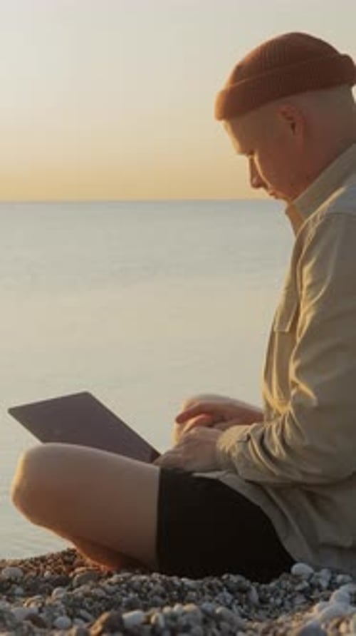 Side View of Man Working on Laptop While Sitting By the Sea at Sunrise