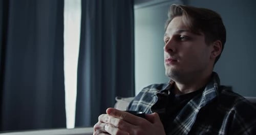 Sad Guy with a Lifeless Look Sits on the Sofa and Drinks a Hot Drink at Home