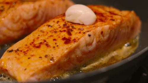 Close Up of Salmon Fish Fillet Cooking in Frying Pan with Oil and Butter with Garlic Resting on Top