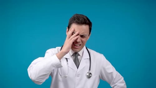 Exhausted Male Doctor Touching Forehead in Studio