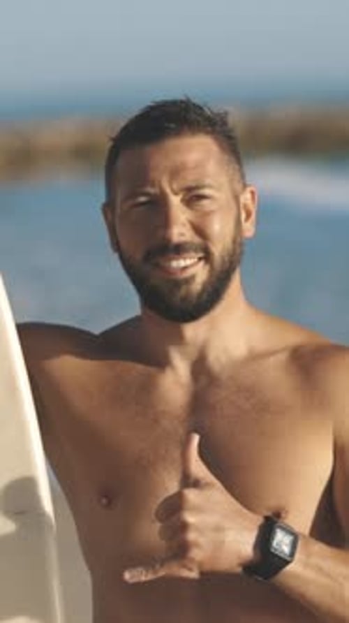 Smiling Man Holding Surfboard Giving Shaka Sign on Beach