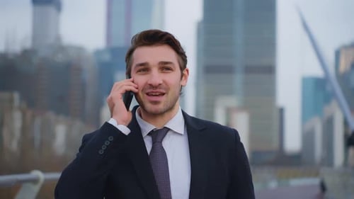 Businessman in suit talking on mobile phone while walking outdoors with city skyscrapers background