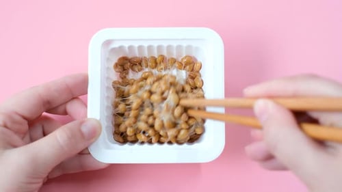 Female hand holding Japanese natto beans with wooden chopsticks on pink background, top view.