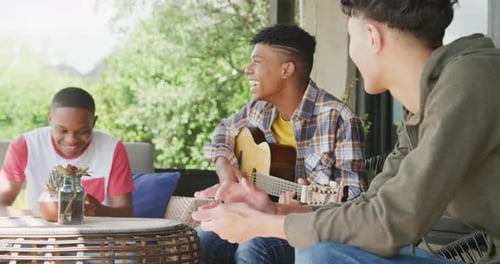 Happy diverse male teenage friends playing guitar at home, slow motion