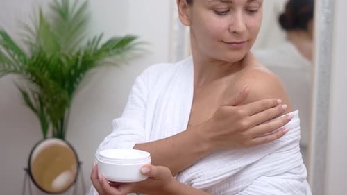 Positive Young Woman Applying Moisturizing Cream on Bare Shoulder in Bathroom Body Care